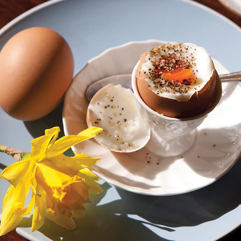 Hard-boiled egg with a soft-boiled yolk with Seagreens Mineral Salt on a white plate, accompanied by a yellow flower on a blue surface.