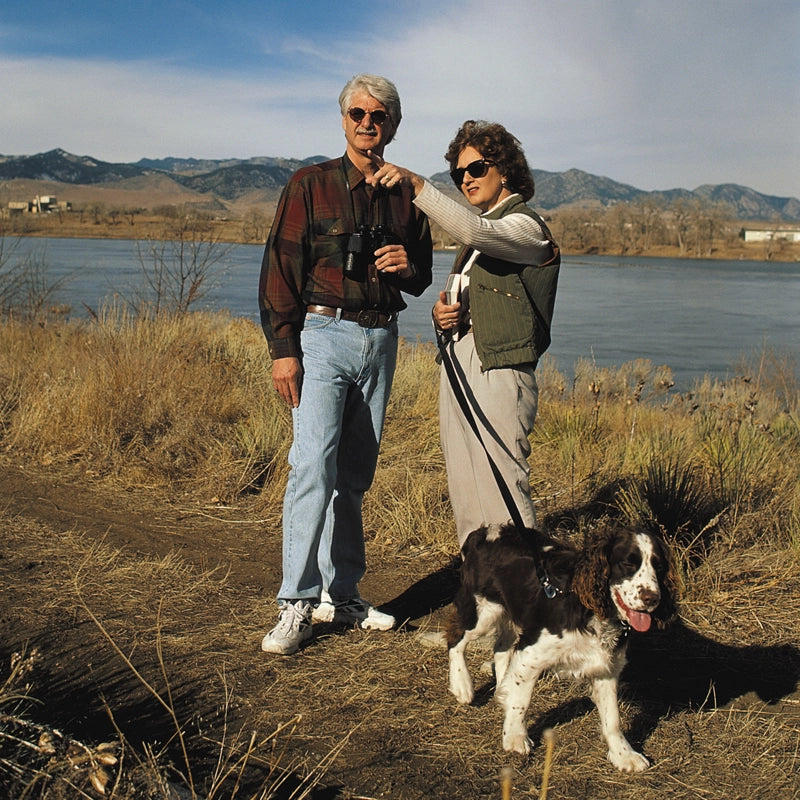 Two people with a dog by a lake with mountains in the background